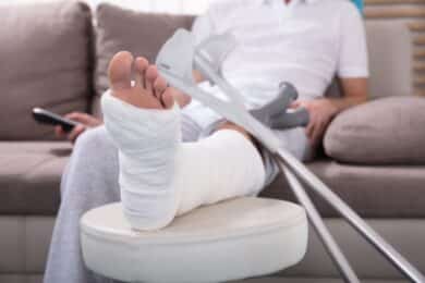 Young Man With Broken Leg Sitting On Sofa Holding Remote